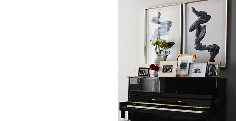“It’s so important to incorporate what people love into their space,” says Lainey. To celebrate Jenny’s passion for playing the piano, Lainey created a focal wall around the piano. “The apartment has very high ceilings so we designed up above the piano in order to draw the eye in.” The framed photos make personal mementos prominent and the black abstract art echoes the color of the piano itself.
