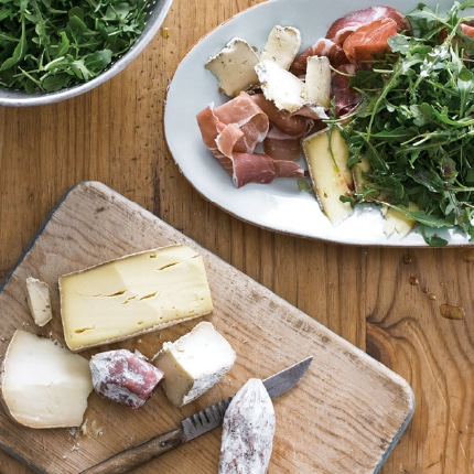 A white serving platter piled high with prosciutto, coppa, a variety of cheeses and arugula.