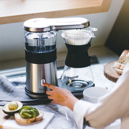 A woman turning on Brim eight-cup automatic pour over coffee maker with avocado toast.