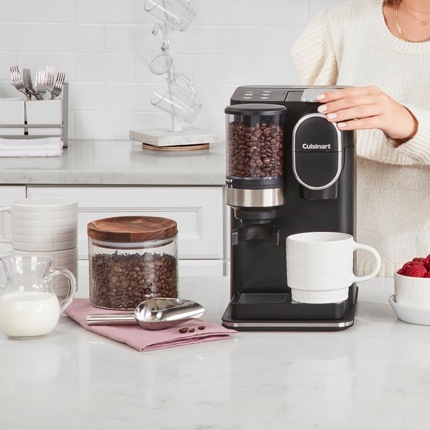 Cuisinart single serve grind and go coffee maker on a counter next to a canister of coffee beans, a glass of milk and a bowl of strawberries.