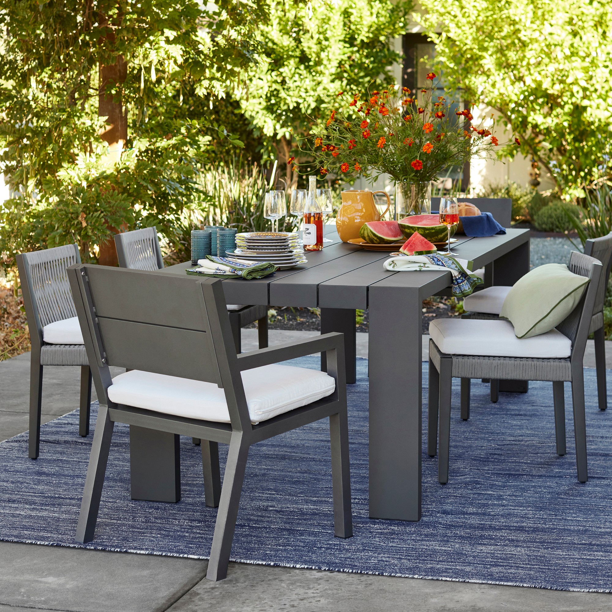 Outdoor dining table set with six chairs and a vase of orange flowers on a slate-gray table, set for a meal with plates, napkins and fruit centerpiece.