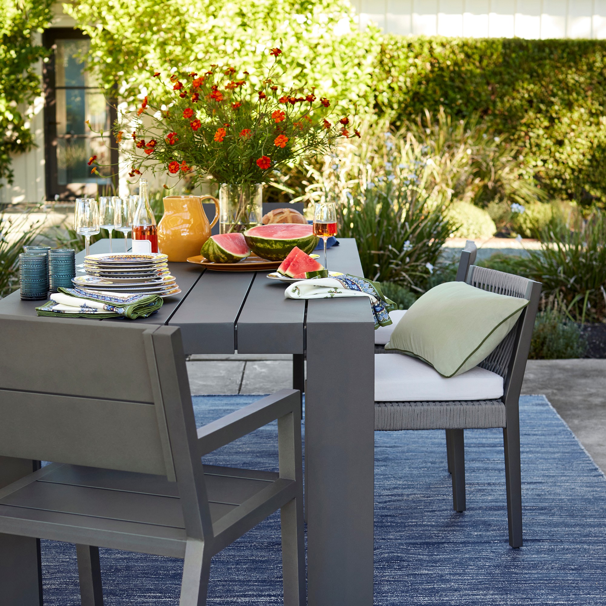 Outdoor dining table with plates, glasses, napkins, watermelon slices, a pitcher and a vase of red flowers.