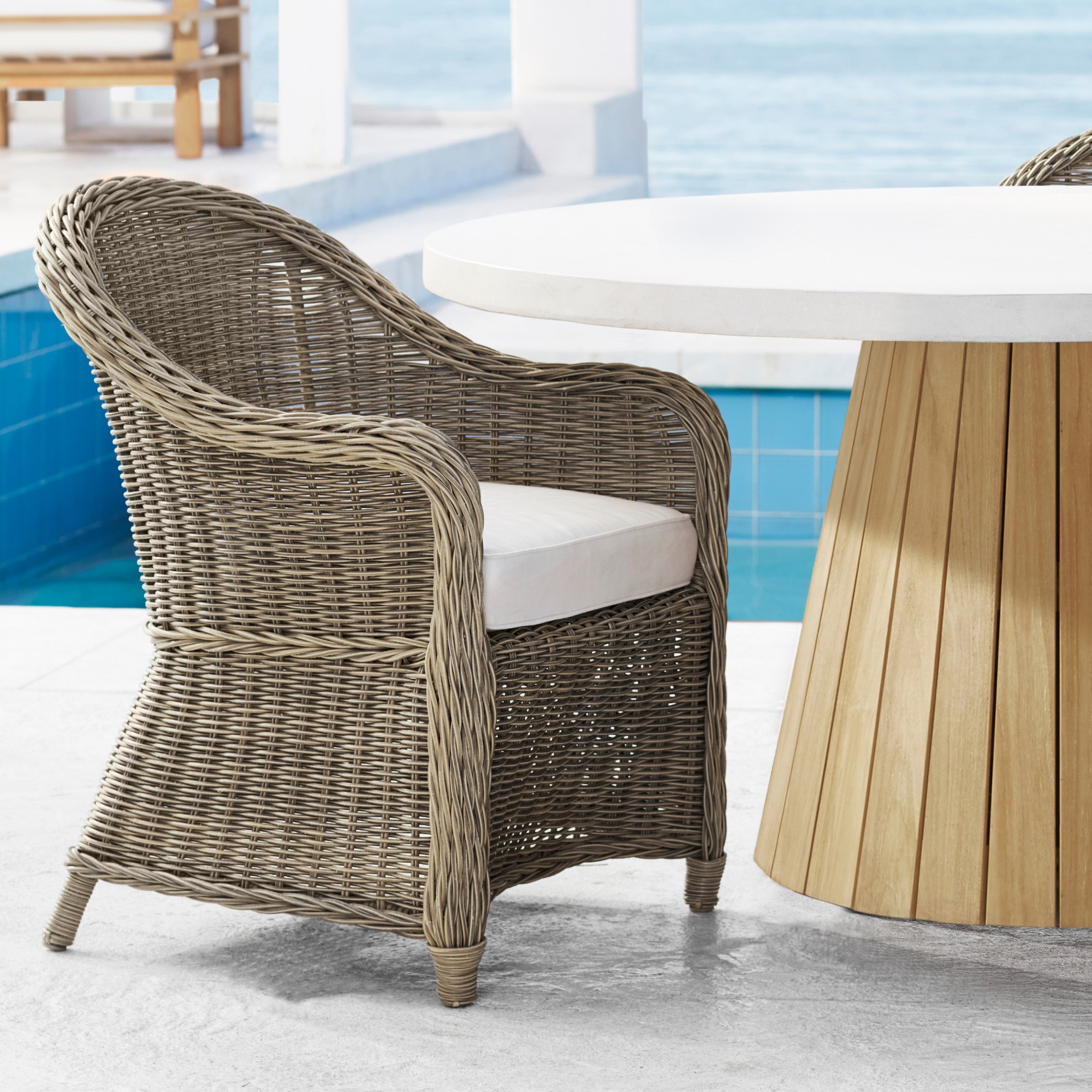 A woven rattan outdoor chair with a white cushion beside a round white table by a pool