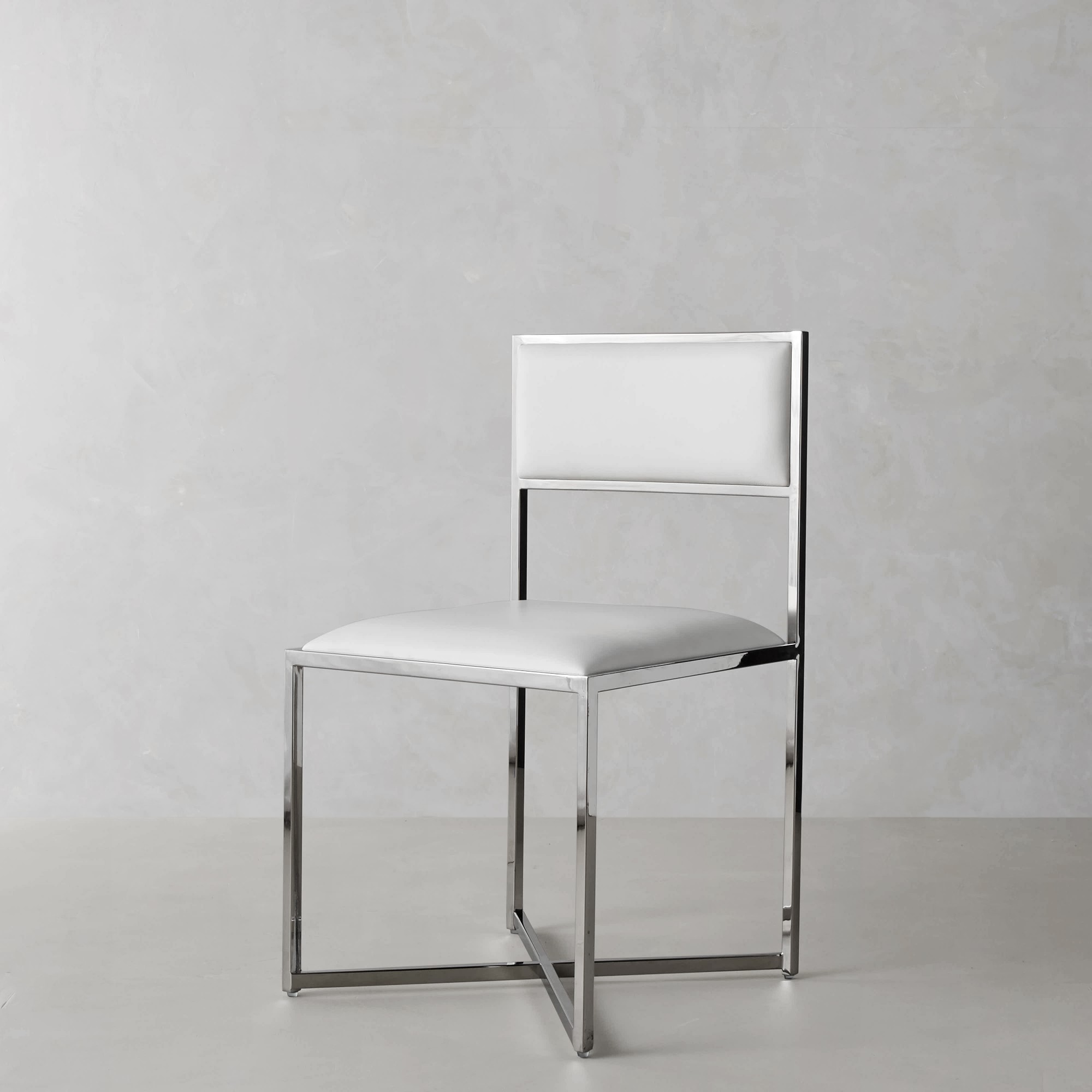 Minimalist white chair with chrome frame and a white upholstered seat and backrest, photographed in a studio against a gray textured wall.