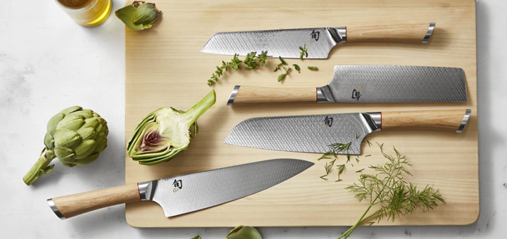 Santoku knife with collection of Japanese-style kitchen knives on a wooden cutting board with herbs, oil and artichoke.