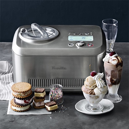  Several types of ice cream and stacks of ice cream sandwiches with a jar of sprinkles arranged in front of a Breville ice cream maker.