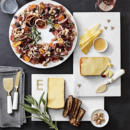 Various types of cheese on a marble and brass monogrammed cheese board with the letter E arranged with another marble cheese board and a platter of food.