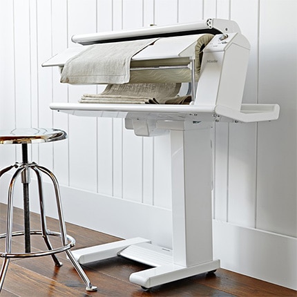 Linens being pressed by a Miele rotary iron next to a metal stool.