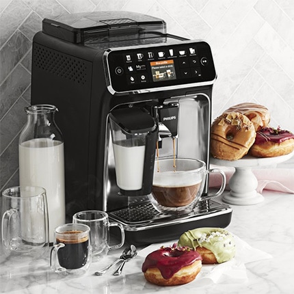A latte being poured from a Philips automatic espresso maker next to plates of donuts and double walled glass mugs.