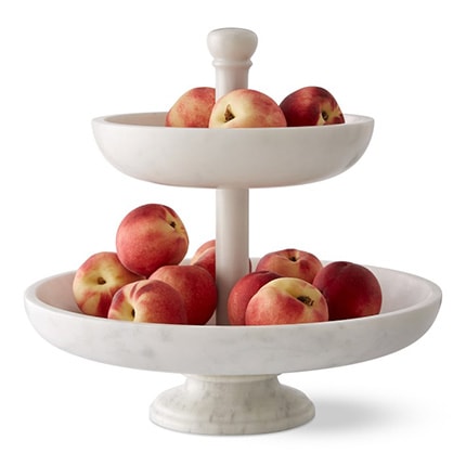 Peaches displayed in a two-tiered white marble fruit bowl.