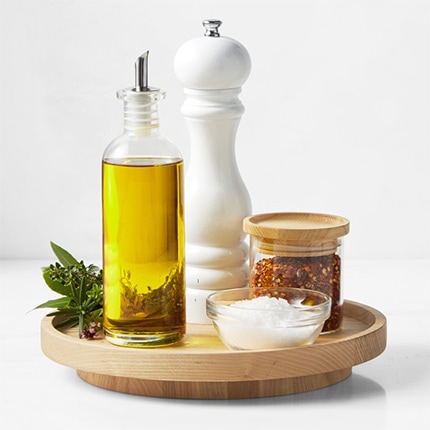 A small wooden Hold Everything lazy Susan holding a flower, bottle of olive oil, red pepper flakes, salt, and pepper grinder.