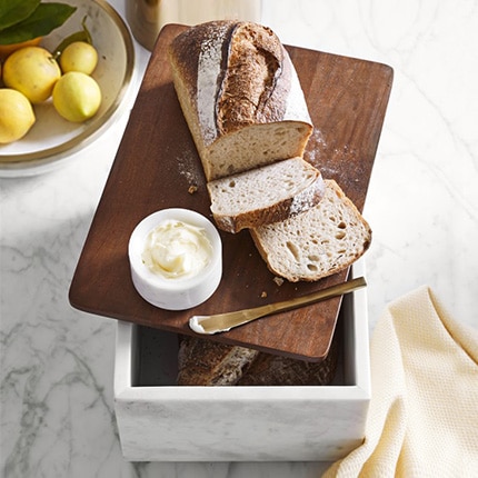 Sliced bread with butter and a butter knife on top of a marble bread box next to a bowl of lemons on a counter.