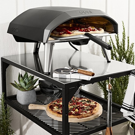  A fresh pizza on a platter on a shelf below an Ooni pizza oven with various utensils.