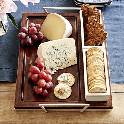 Wooden cheese tray with various cheeses, crackers and grapes on a table with a blue tablecloth and vase with flowers.