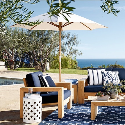 A swimming pool next to Larnaca teak umbrella and several pieces of outdoor furniture with trees and the ocean in the background.