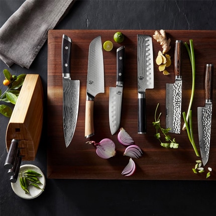 An array of nakiri and santoku knives on a wooden cutting board with onion, lime, ginger, chives and herbs.