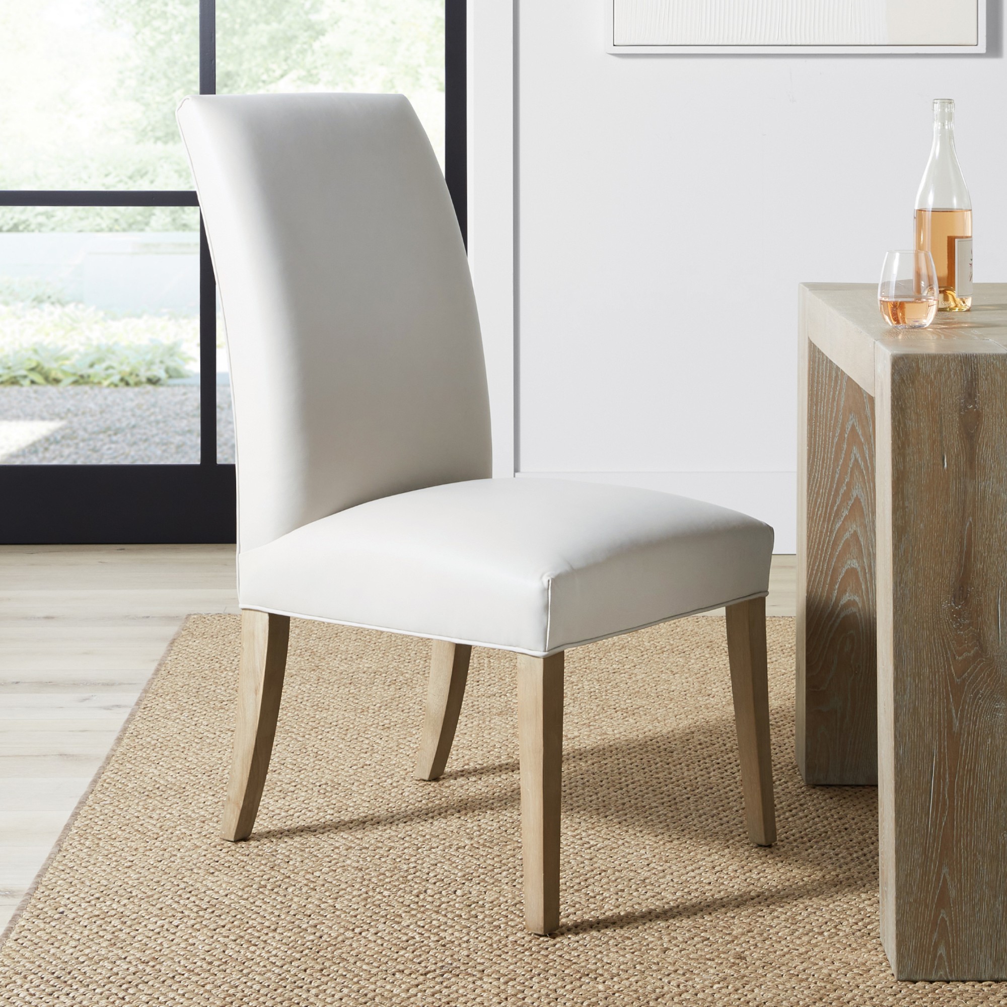 Beige upholstered dining chair with light wooden legs, placed indoors near a bottle and glass.