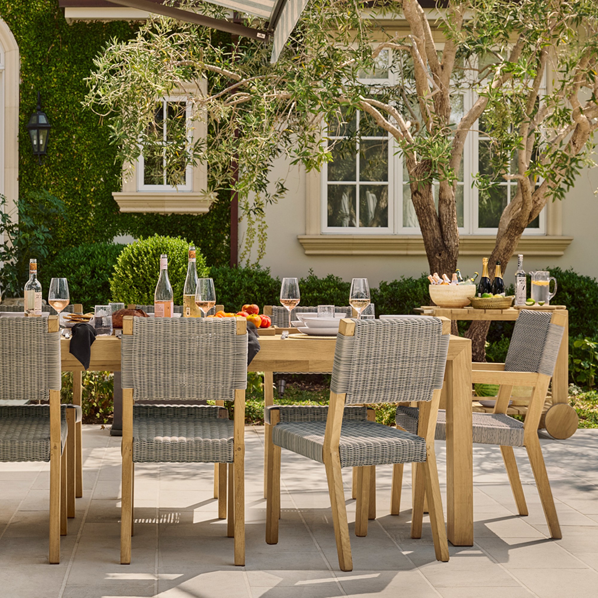 Outdoor dining table set with woven chairs arranged for a meal on a patio, bottles and fruit on table